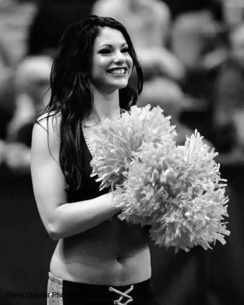 Black-and-white portrait of Kelsey in a cheer uniform, smiling and holding pom-poms