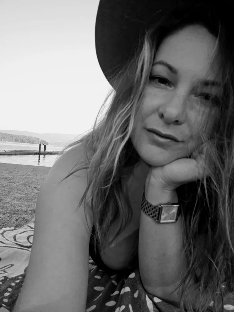 Black-and-white selfie of Kelsey in a wide-brimmed hat, lying on a beach by a lake