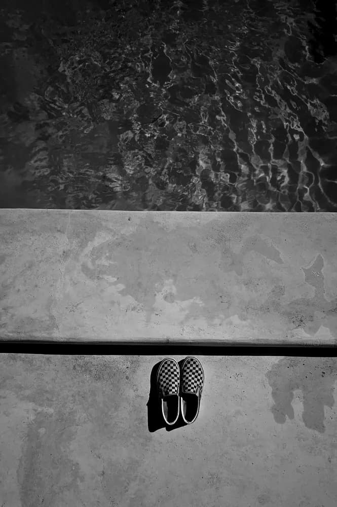 Black-and-white overhead shot of checkered slip-on shoes on a pool's concrete edge