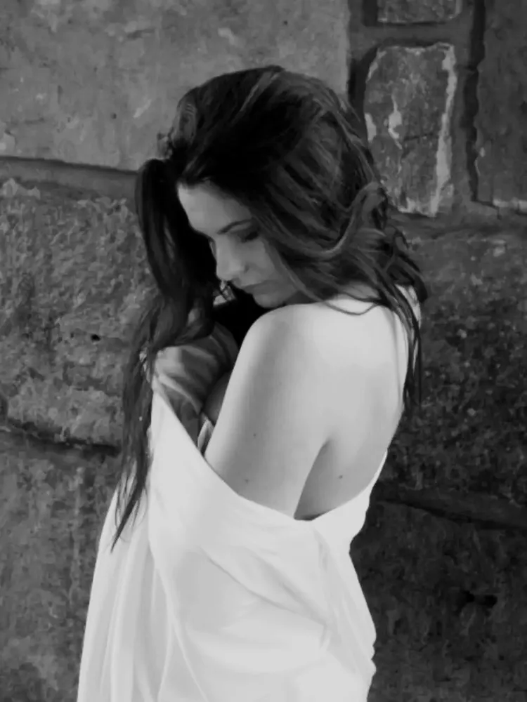 Black-and-white photo of Kelsey against a stone wall, wrapped in white fabric and looking down over one shoulder