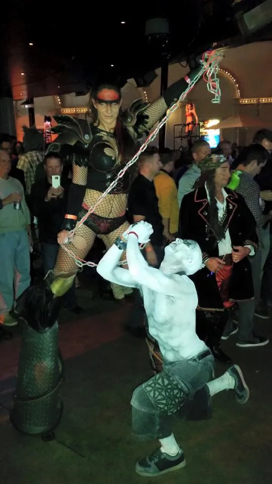 A winged warrior costume on stilts leading a performer in white body paint on a chain