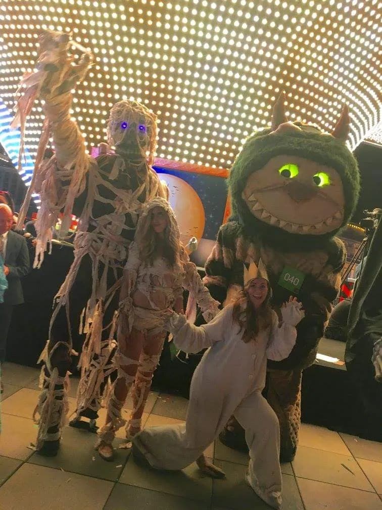A mummy on stilts beside a Wild Things costume under a wall of lights